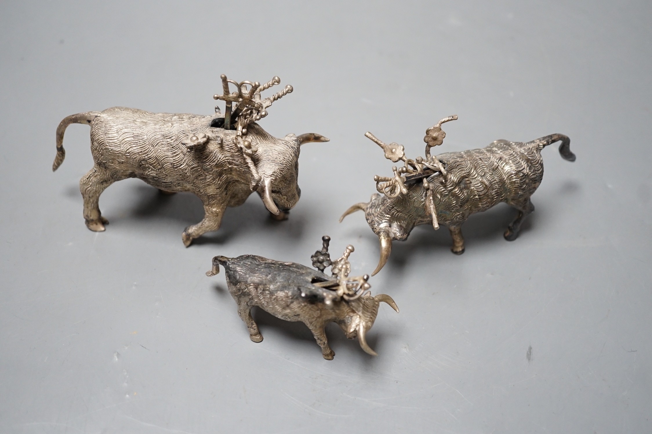Three miniature white metal model cocktail stick holders? each modelled as a bull with sword shaped sticks, two stamped sterling 925, largest length 12.3cm.